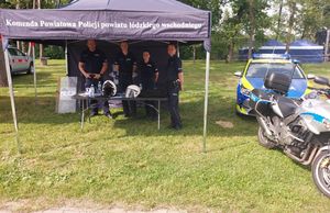 Czterech policjantów stoi pod granatowym namiotem na którym jest napis Komenda Powiatowa Policji powiatu Łódzkiego Wschodniego. Obok namiotu stoi policyjny radiowóz i motocykl.