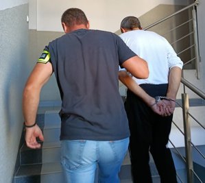 Policjant z opaską na ramieniu z napisem policja prowadzi w górę po schodach zatrzymanego mężczyznę