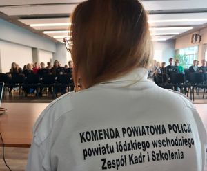 Kobieta w białej bluzce siedzi odwrócona tyłem przed dużą grupą osób, na plecach ma napis Zespół kadr i szkolenia Komendy Powiatowej Policji powiatu łódzkiego wschodniego.