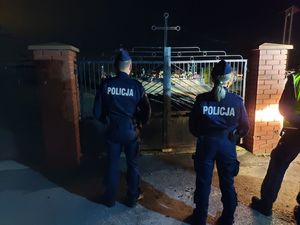 Dwóch policjantów i policjantka w środku stoją przed bramą cmentarza.