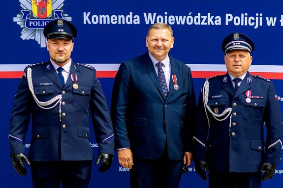 Dwóch policjantów a po środku mężczyzna w garniturze stoją obok siebie na ściance z napisem Komenda Wojewódzka Policji w Łodzi.