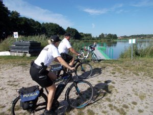 Dwóch policjantów na rowerach patroluje miejsce przy zbiorniku wodnym.