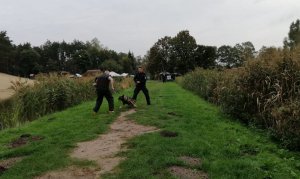 Pozorant udający napastnika jest przeszukiwany przez policjanta i jednocześnie obserwowany przez psa, który stoi przed nim.
