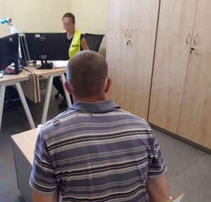 Policjantka ubrana w żółtą kamizelkę z napisem policja siedzi za biurkiem i przesłuchuje zatrzymanego. Mężczyzna siedzi na wprost policjantki.