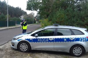 Policjant umundurowany z żółtą kamizelką z napisem policja stoi przy drodze obok radiowozu oznakowanego i mierzy prędkość.