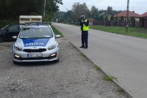 Policjant umundurowany z żółtą kamizelką z napisem policja stoi przy drodze obok radiowozu oznakowanego i mierzy prędkość.