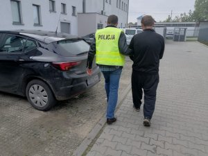 Policjant nieumundurowany, który ma założoną żółtą kamizelkę z napisem policja prowadzi obok idącego zatrzymanego mężczyznę do samochodu.