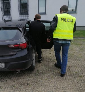Policjant nieumundurowany, który ma założoną żółtą kamizelkę z napisem policja prowadzi obok idącego zatrzymanego mężczyznę do samochodu.