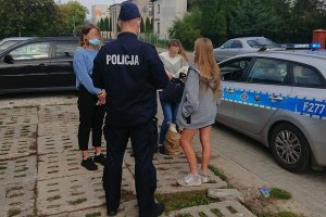 Policjanci w mundurze podjęli interwencję wobec osób nie mających założonych maseczek na twarzy. Policjanci stoją przy radiowozie i legitymują stojące obok nich osoby.