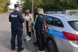 Policjanci w mundurze podjęli interwencję wobec osób nie mających założonych maseczek na twarzy. Policjanci stoją przy radiowozie i legitymują stojące obok nich osoby.