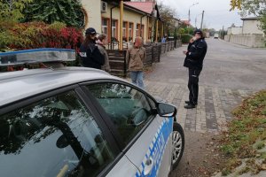 Policjanci w mundurze podjęli interwencję wobec osób nie mających założonych maseczek na twarzy. Policjanci stoją przy radiowozie i legitymują stojące obok nich osoby.