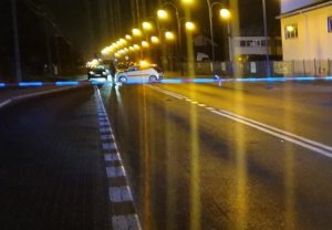 ulica odgrodzona taśmą z napisem policja gdzie doszło do wypadku śmiertelnego, widać w oddali stojący na jezdni samochód i radiowóz z włączonymi sygnałami świetlnymi