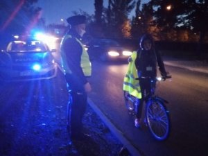 Umundurowany policjant w żółtej kamizelce odblaskowej stoi przed radiowozem gdzie są włączone sygnały świetlne a na wprost policjanta stoi kobieta przy rowerze i zakłada kamizelkę