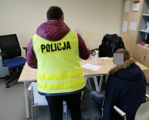 Nieumundurowany policjant stoi tyłem, ma założoną żółtą kamizelkę z napisem policja, obok niego siedzi na krzesełku zatrzymany mężczyzna