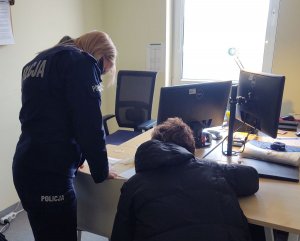 Umundurowana policjantka stoi tyłem przy zatrzymanej kobiecie, która siedzi tyłem przy biurku