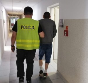 Policjant w żółtej kamizelce z napisem policja prowadzi zatrzymanego mężczyznę, obaj są odwróceni plecami