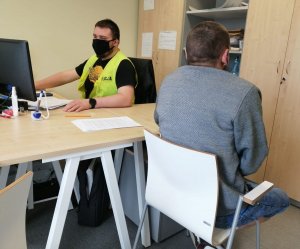 policjant w żółtej kamizelce z napisem policja siedzi przy biurku przed komputerem, na twarzy ma założoną czarną maseczkę ochronną. Zatrzymany mężczyzna siedzi bokiem do biurka a przodem do policjanta