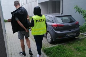Policjantka w żółteju kamizelce z napisem policja prowadzi zatrzymanego mężczyznę, obok stoi samochód