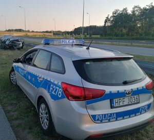 Policyjny radiowóz stoi na poboczu przy rozbitym samochodzie.