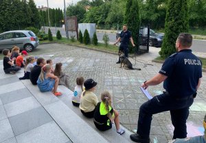 umundurowani policjanci stoją przed dziećmi siedzącymi na schodkach, obok jednego z policjantów siedzi pies