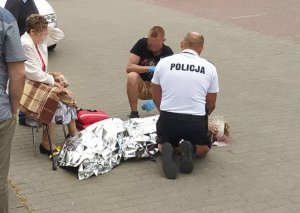 umundurowany policjant udziela pomocy leżącej kobiecie, policjant jest schylony przy kobiecie wraz z innym mężczyzną który mu pomaga. Obok siedzi na krzesełku inna kobieta i stoi dwóch mężczyzn