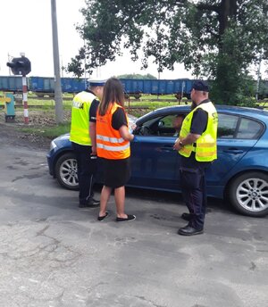 Umundurowani policjanci i pracownicy sok podchodzą do samochodów stojących przed przejazdem kolejowym.