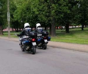 Dwaj umundurowani policjanci , w czarnych kurtkach z napisem na plecach Policja i w białych kaskach jadą drogą na motocyklach