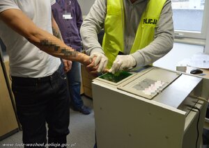 Nieumundurowany policjant trzyma aparat fotograficzny i fotografuje zatrzymanego oraz pobiera jego odciski palców.