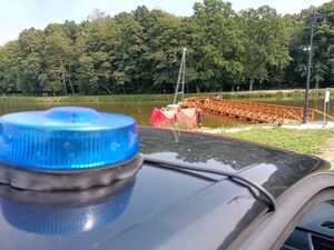 Na fotografii widoczny dach stojącego nieoznakowanego radiowozu, na którym przymocowany jest niebieski sygnalizator w oddali widać parawan strażacki koloru czerwonego i stojących przed nim strażaków umundurowanych i policjantów nieumundurowanych odwróconych tyłem.