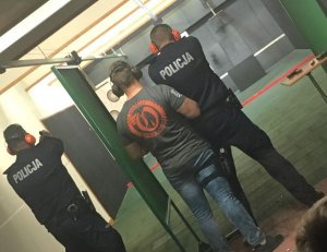 Policjanci w cywilnych ubraniach stoją obok umundurowanych policjantów, wszyscy mają nauszniki na uszach i są odwróceni tyłem. Stoją na stanowiskach przygotowanych do strzelania.