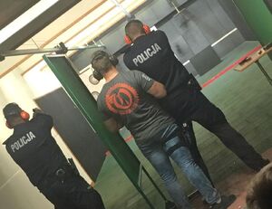 Policjanci w cywilnych ubraniach stoją obok umundurowanych policjantów, wszyscy mają nauszniki na uszach i są odwróceni tyłem. Stoją na stanowiskach przygotowanych do strzelania.