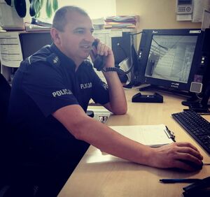 Policjant w granatowym mundurze rozmawia przez telefon, siedzi przy biurku na którym ustawione są komputery.