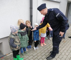 umundurowany policjant wyciąga rękę do dzieci stojących w rzędzie pod ścianą