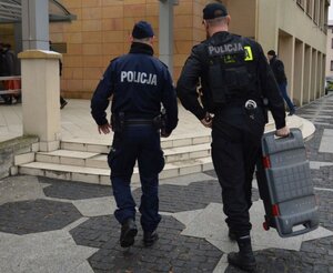 Umundurowani policjanci wchodzą do budynku, jeden z nich trzyma walizkę.