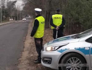 dwóch umundurowanych policjantów w żółtych kamizelkach z napisem policja stoi przy radiowozie zaparkowanym przy drodze