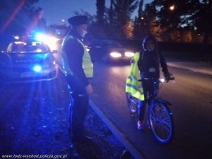 umundurowany policjant w żółtej kamizelce stoi przed radiowozem w którym migają niebieskie światła, policjant patrzy w kierunku rowerzystki, która stoi przed nim prowadząc rower
