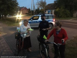 umundurowana policjantka stoi przy dwóch rowerzystkach, które trzymają kamizelki odblaskowe, za nimi stoi zaparkowany radiowóz