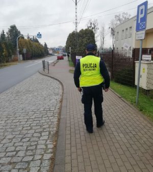 policjant umundurowany w żółtej kamizelce z napisem Policja dzielnicowy stoi odwrócony tyłem na chodniku przed szkołą