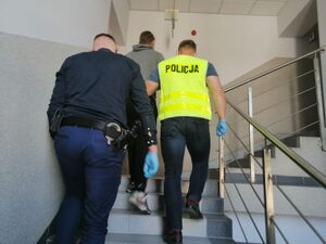 Dwóch policjantów,jeden z nich w żółtej kamizelce z napisem policja wchodzi po schodach, w środku nich idzie zatrzymany mężczyzna.