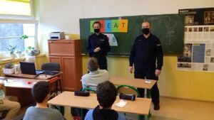 Policjantka i policjant w granatowych mundurach i w maseczkach na twarzy stoją przed uczniami siedzącymi w ławkach.