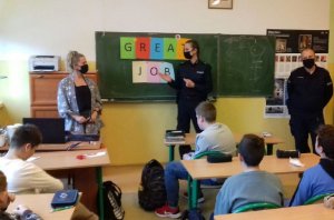 Policjantka i policjant w granatowych mundurach i w maseczkach na twarzy stoją przed uczniami siedzącymi w ławkach.