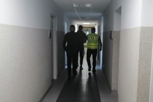 dwóch policjantów prowadzi w środku zatrzymanego mężczyznę, idą korytarzem