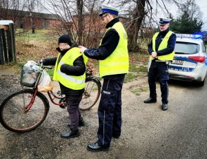 Dwaj umundurowani policjanci w żółtych kamizelkach i białych czapkach na głowie dawali rowerzystom kamizelki odblaskowe i pomagali im je założyć