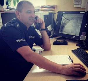 umundurowany policjant siedzi za biurkiem na którym są monitory komputerów i rozmawia przez telefon