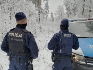 dwóch umundurowanych policjantów stoi przy radiowozie, są odwróceni tyłem, wokółl nich zaśnieżone drzewa i droga