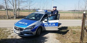 Policjant stoi przy otwartych drzwiach radiowozu i rozmawia przez telefon z osobą przebywającą na kwarantannie.