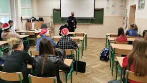 Policjant w niebieskim mundurze stoi na środku klasy i mówi do młodych ludzi siedzących przed nim w ławkach.