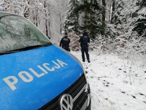 dwóch policjantów stoi odwróconych tyłem, wokół są drzewa pokryte śniegiem w pobliżu stoi zaparkowany radiowóz