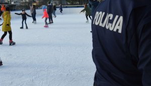 mundur policjanta odwróconego tyłem, na granatowym mundurze biały napis Policja, w tle widać dzieci jeżdżące na łyżwach na lodowisku