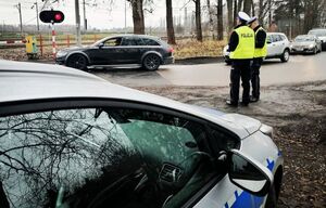 Dwóch policjantów w białych czapkach i żółtych kamizelkach stoi obok drogi na której ustawione są w rzędzie samochody stojące przy opuszczonym szlabanie kolejowym, za policjantami stoi zaparkowany radiowóz.